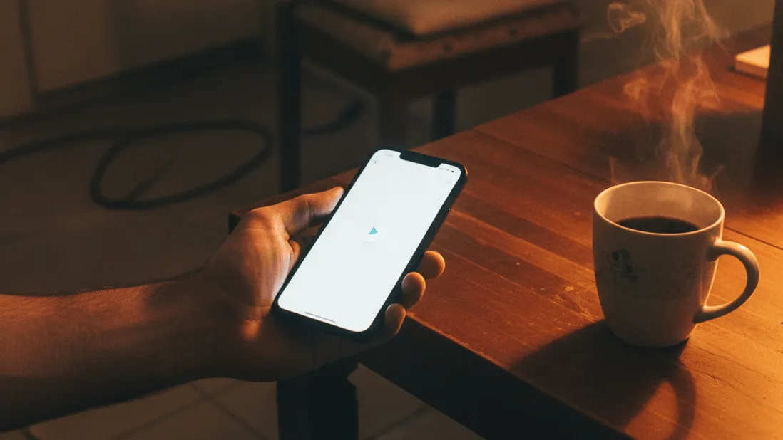 A hand holds a smartphone showing a blank paused video app screen above a kitchen table with a steaming coffee mug.