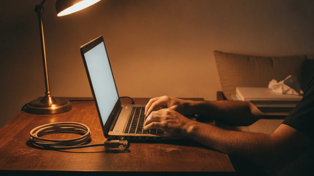 A man's hands type on a laptop keyboard at night, illuminated by a desk lamp and screen glow.