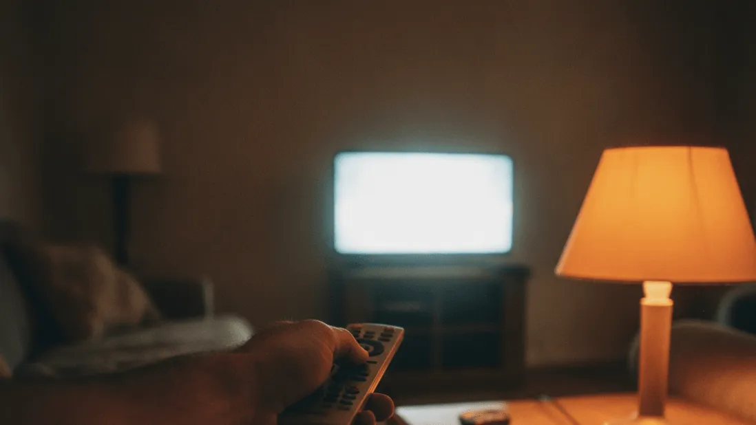 A hand holds a remote toward a TV screen, a lamp illuminates a notepad on a side table.