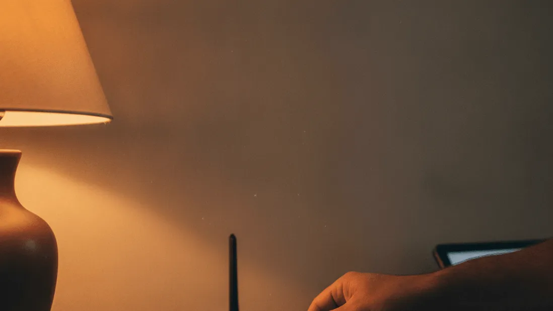 A man's hand connects an ethernet cable to a router on a wooden shelf in warm evening light.