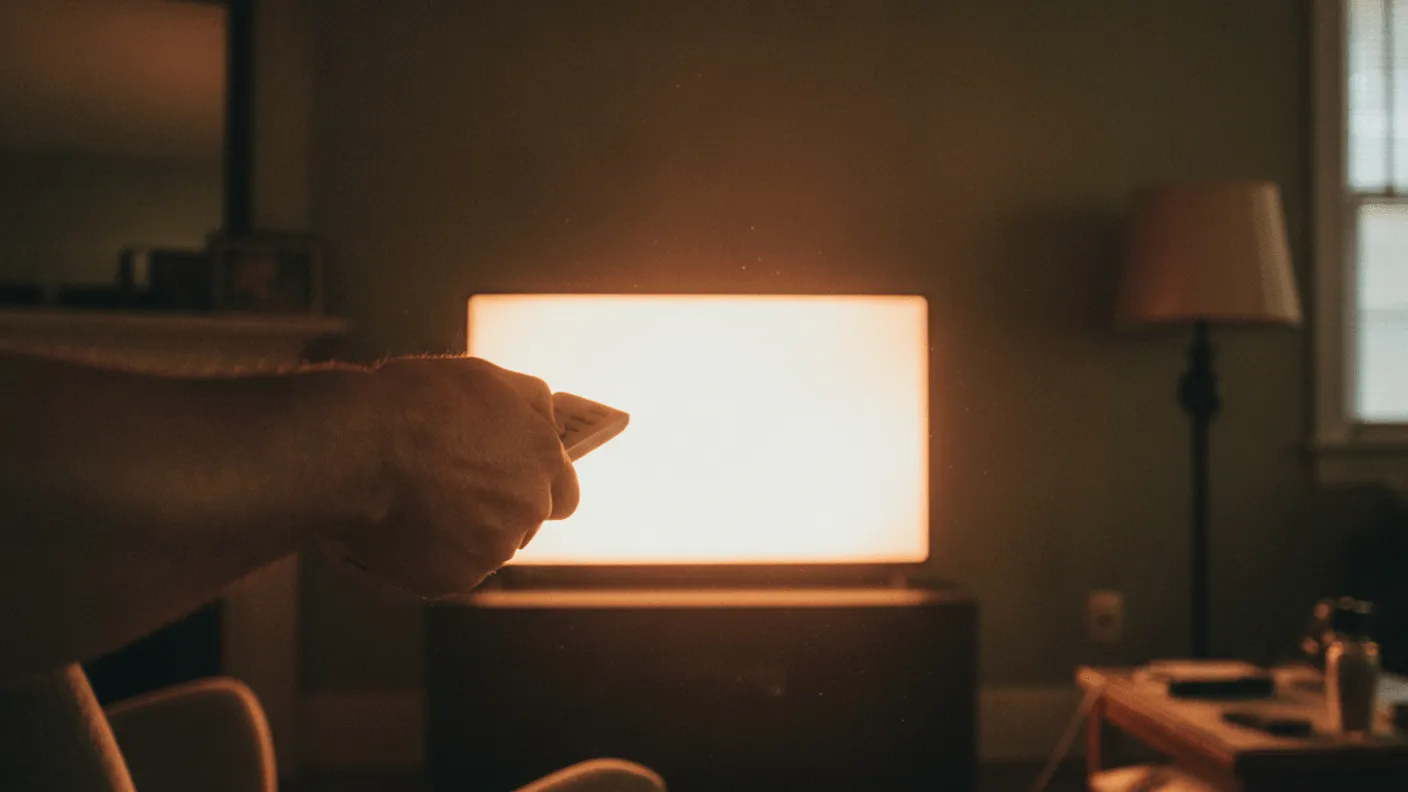 A man's hand holds a remote, pointing at a softly glowing television screen in a dark, warm-lit living room.