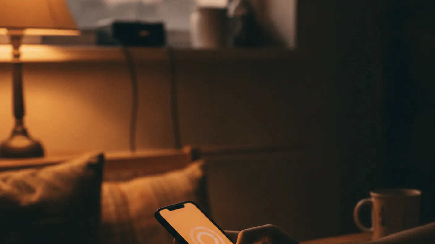 A hand holds a smartphone showing a loading spinner above a kitchen table at dusk, a sticky note and a pen beside it.