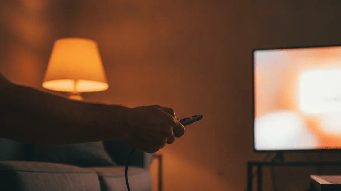 A man's hand holds a remote, an HDMI cable lies on the floor, and a warm lamp illuminates the room.