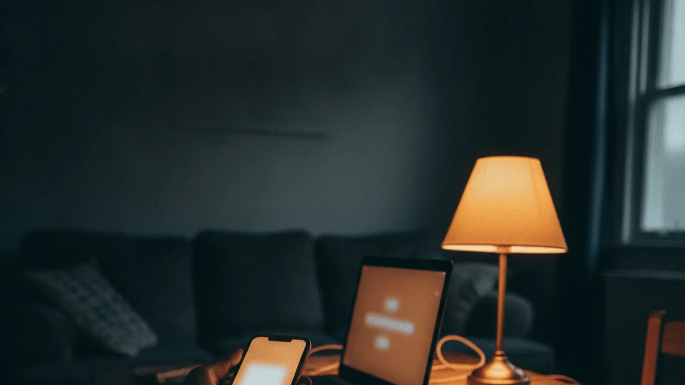 A hand holds a phone with a bright screen at a wooden kitchen table, next to a laptop and notepad in low light.